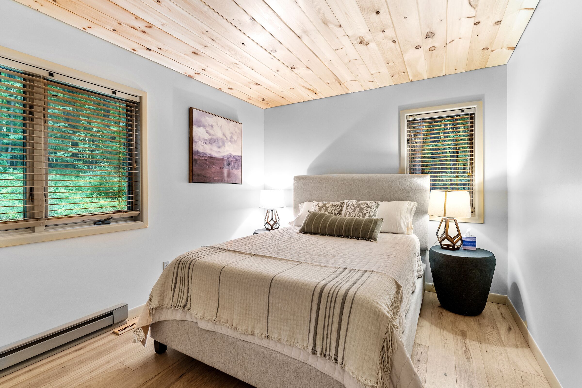 Ein Schlafzimmer mit Holzbalkendecke, zwei Fenstern mit Jalousien, einem Bett mit beiger Decke, einem Nachttisch mit Lampe und einem Bild an der Wand.