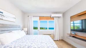 A bedroom with a large bed, a TV on a wooden stand, and a view of the sea through the window. - Beautiful Oceanfront  2 bedroom, 2 bath condo Aguada, PR (Aguada)