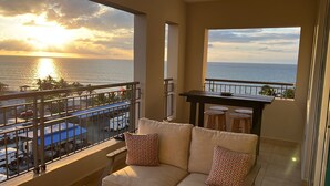A balcony with a view of the sea at sunset, a sofa with patterned cushions, and a small table with stools. - Beautiful Oceanfront  2 bedroom, 2 bath condo Aguada, PR (Aguada)