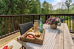 Een houten terras met een rieten stoel, een tafel met een dienblad eten, een fles en een potplant.
