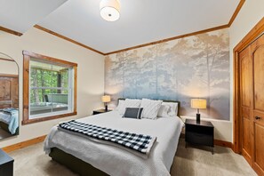 Een slaapkamer met een groot bed, een nachtkastje, een lamp, een kledingkast en een raam met uitzicht op bomen.