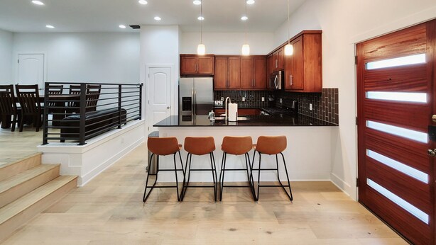 A modern kitchen with wooden cabinets, stainless steel appliances, and a black countertop. There are three bar stools and a dining table with chairs. - Downtown Stay in East Austin @ 12th Street (Austin)