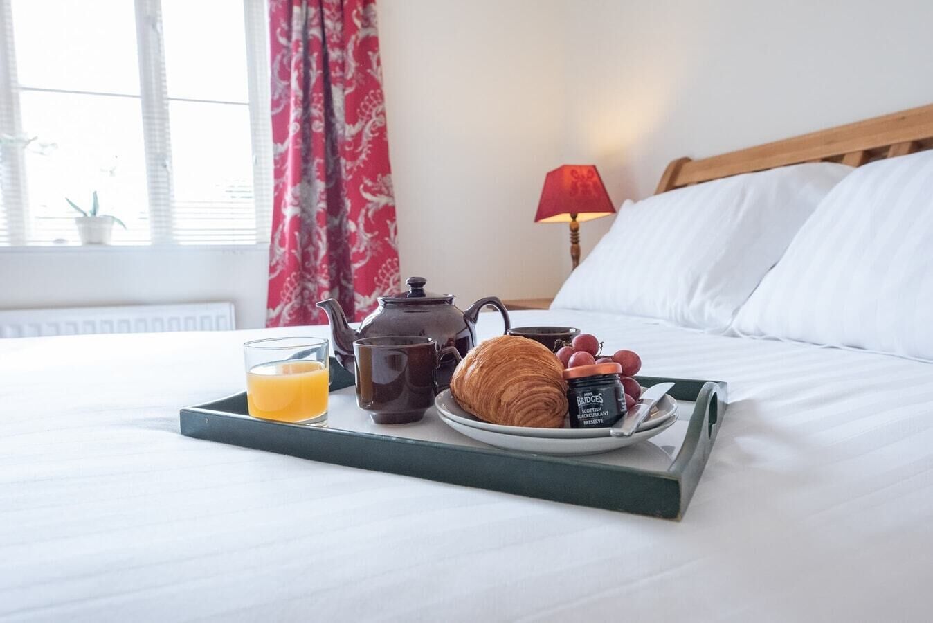 A neatly made bed with white linens, a tray with breakfast items including a croissant, a glass of orange juice, a teapot, and a jar of jam.