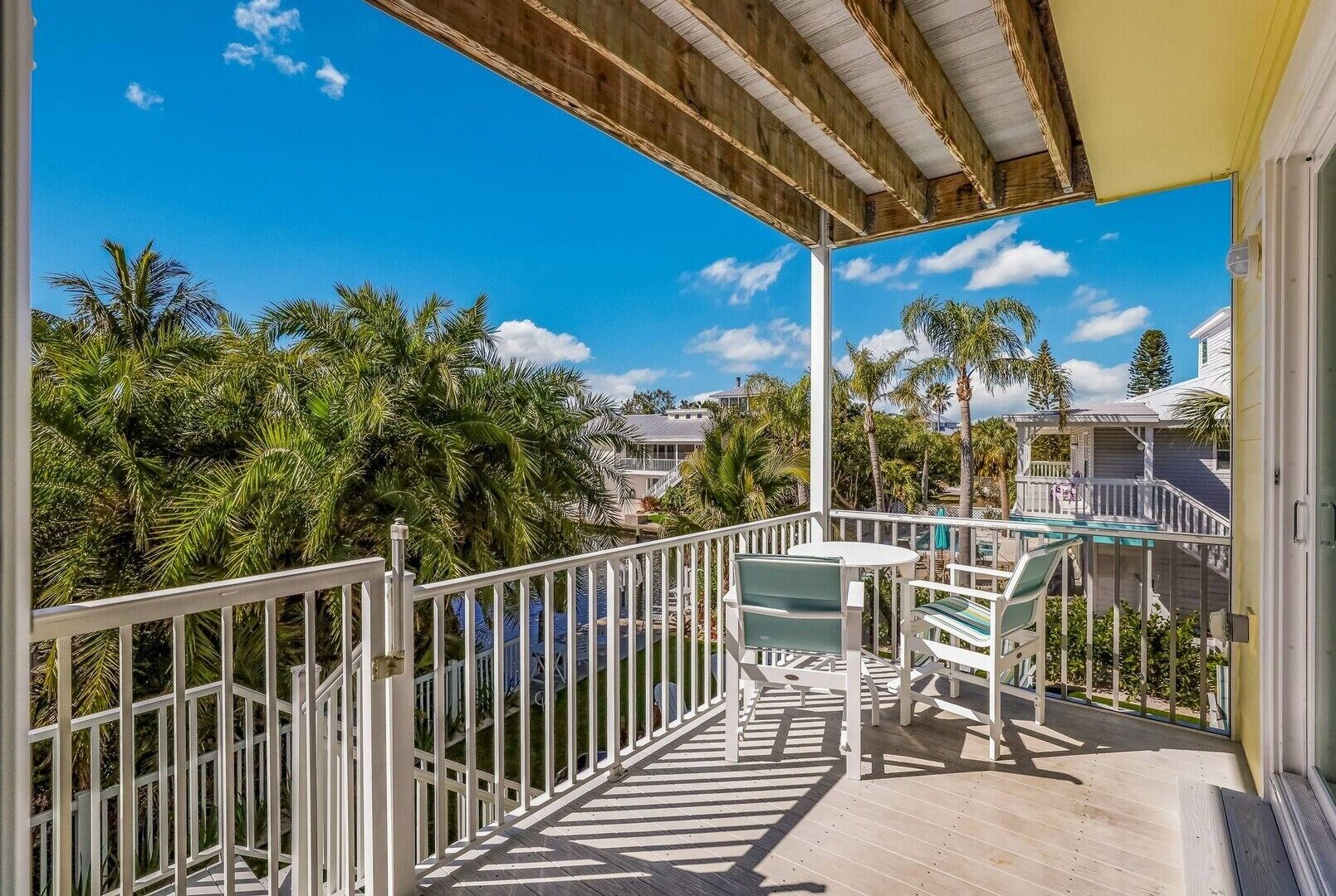 A wooden deck with a white railing, a table with chairs, and a view of palm trees and other buildings.