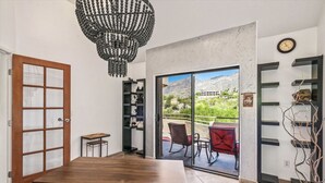 Una habitación con una mesa de comedor de madera, una araña, una estantería, un ventanal corredero que da a un patio exterior con sillas, y un reloj en la pared.