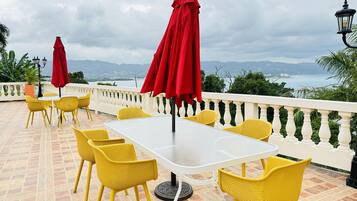 Un patio al aire libre con sillas amarillas y sombrillas rojas, con vistas a un paisaje montañoso y una masa de agua.