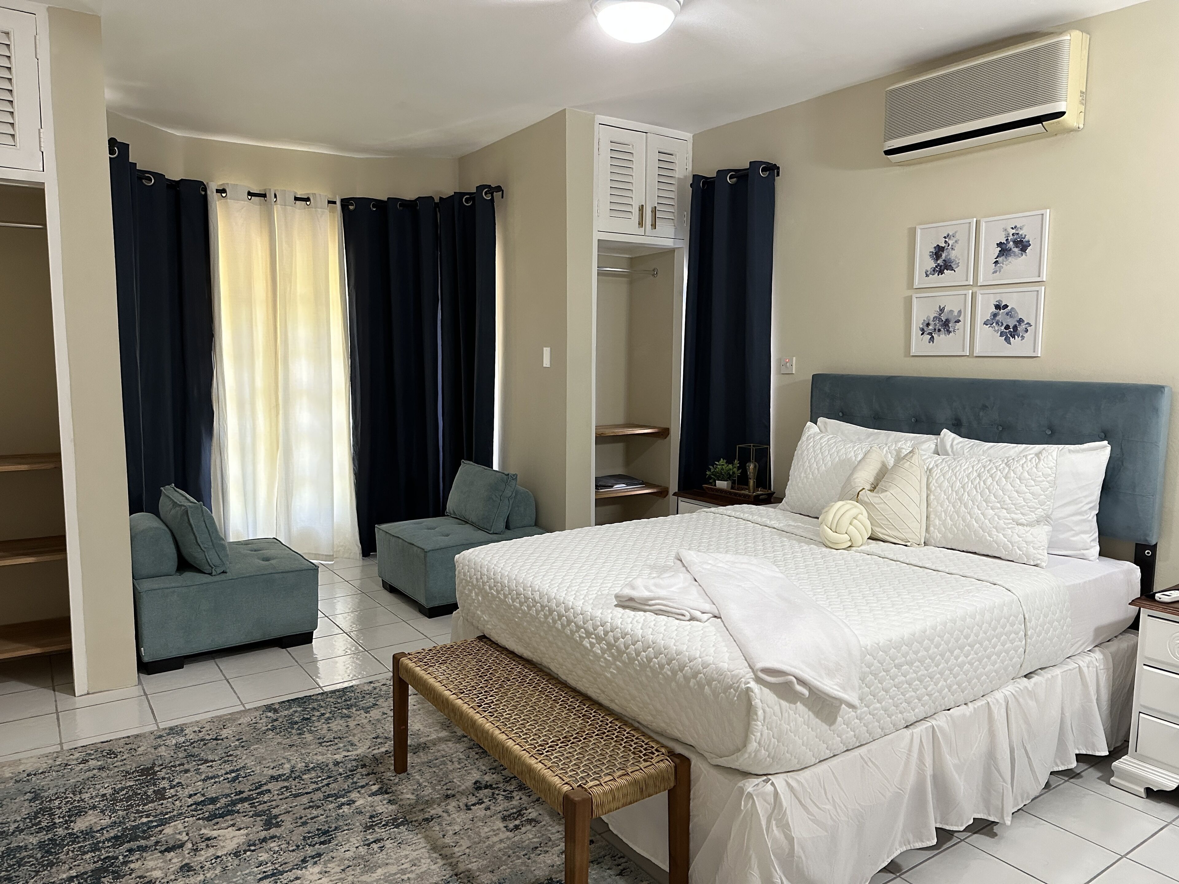 Quarto com cama, mesas de cabeceira, um banco, guarda-roupa e janela com cortinas.