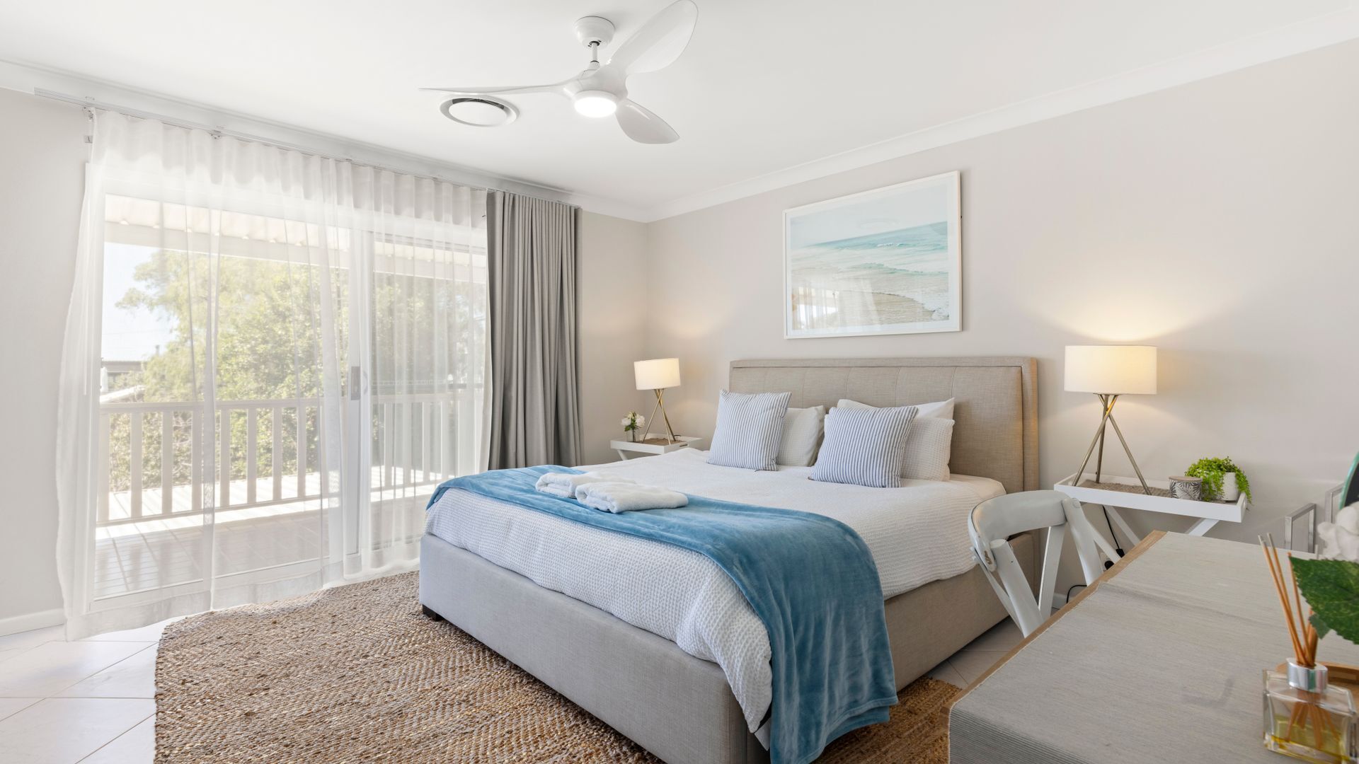 A modern bedroom with a large bed, a ceiling fan, a view of the outdoors, and a bedside table with a lamp.
