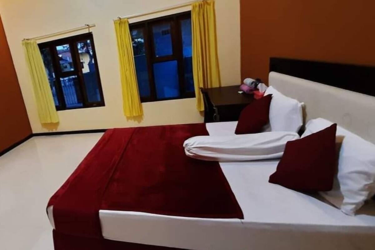 A hotel room with a bed, two windows with curtains, a table with a chair, and an air conditioning unit on the wall.