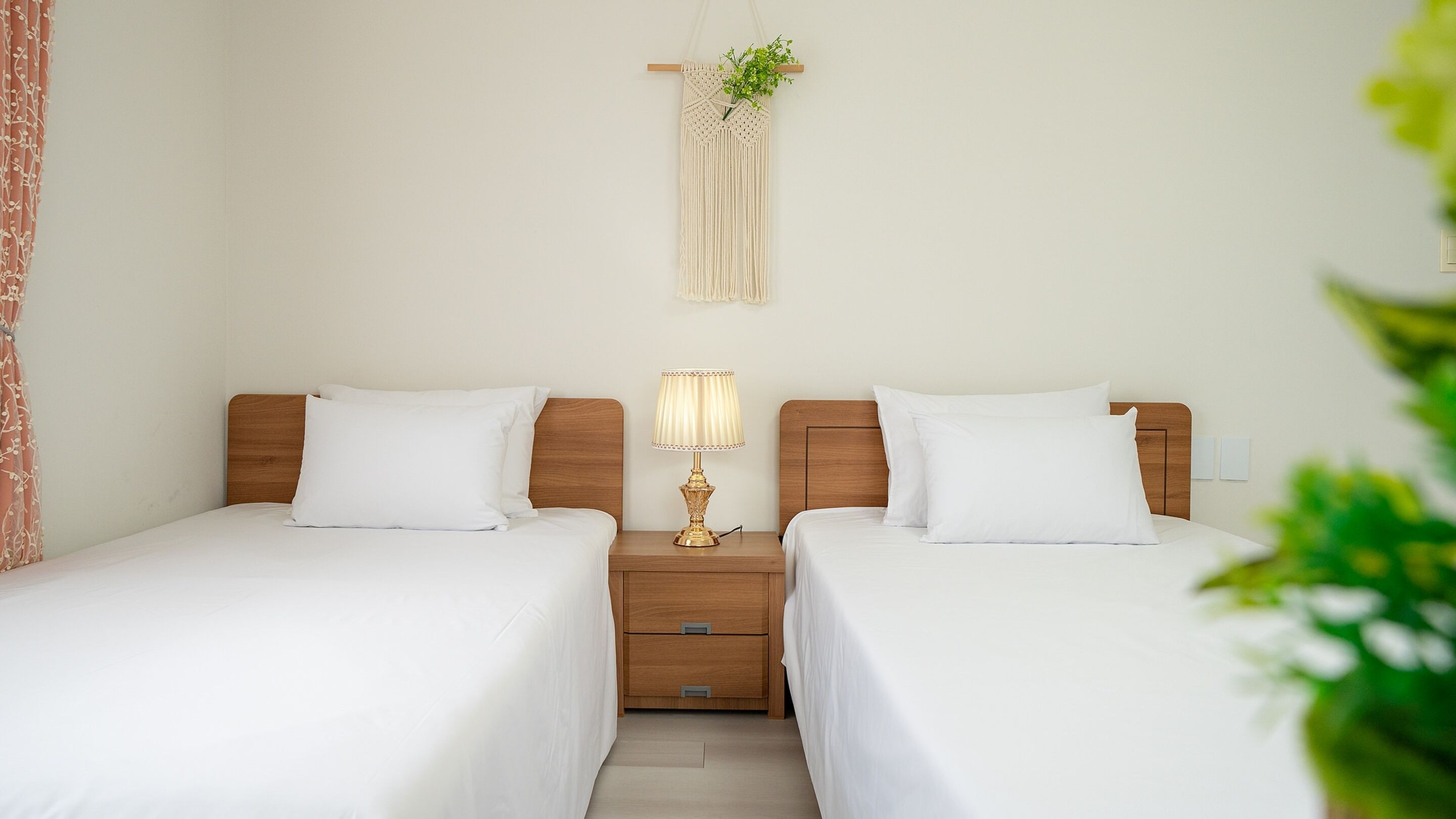 A hotel room with two single beds, a wooden nightstand with a lamp, a macrame wall hanging, and a small plant.
