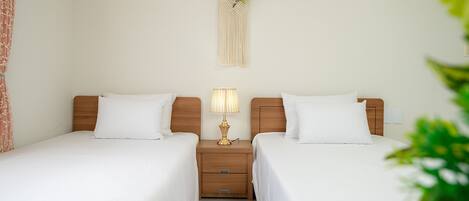 A hotel room with two single beds, a wooden nightstand with a lamp, a macrame wall hanging, and a small plant.