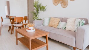 Un salon avec une table basse en bois, un canapé beige agrémenté de coussins à motifs, une table à manger avec des chaises et une grande plante.