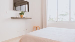 Une chambre à coucher moderne avec un grand lit, un téléviseur fixé au mur, un petit tabouret et une fenêtre avec des rideaux.