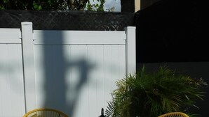 Dos sillas de mimbre amarillas, un farol negro y una planta en maceta en un patio con una cerca blanca y un edificio al fondo.