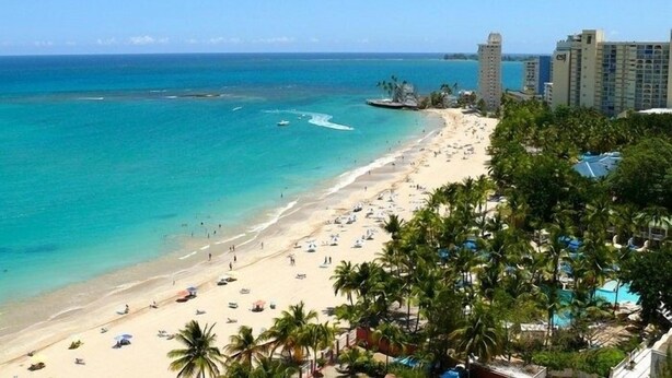 Una playa con aguas turquesas cristalinas, palmeras y edificios altos al fondo.