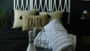 Una cama con cabecera blanca, una almohada con la palabra "FAITH", una botella de champán y dos copas de vino sobre una bandeja.
