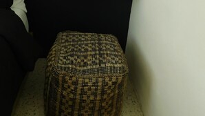 Una habitación pequeña y acogedora con una pared oscura, un estante blanco con libros y velas, un jarrón blanco con una planta y un otoman tejido.