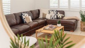 Un canapé d’angle en cuir marron agrémenté de coussins décoratifs, une table basse en fibres tressées avec une plante et des objets décoratifs, et une plante en pot dans un panier tressé.