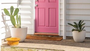 Une maison blanche avec une porte rose, une enseigne indiquant « BURTS », et deux plantes en pot sur le porche.