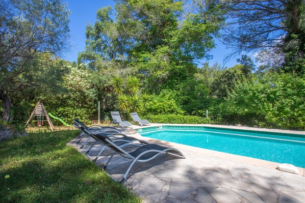 Une piscine avec des chaises longues, une aire de jeux pour enfants et de la verdure tout autour.