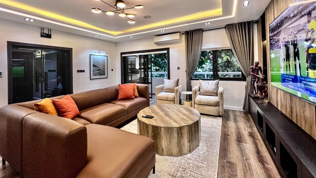 A modern living room with a brown leather sofa, two armchairs, a wooden coffee table, and a flat-screen TV mounted on the wall. - Luxury, contemporary 4-Bed Apartment in Ikoyi (Lagos)