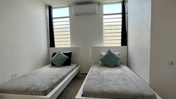 A small, compact room with two single beds, each with a pillow and a headboard. There is an air conditioning unit mounted on the wall above the beds.
