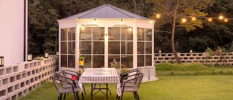 A glass greenhouse with a checkered table and chairs, surrounded by a wooden fence and string lights.
