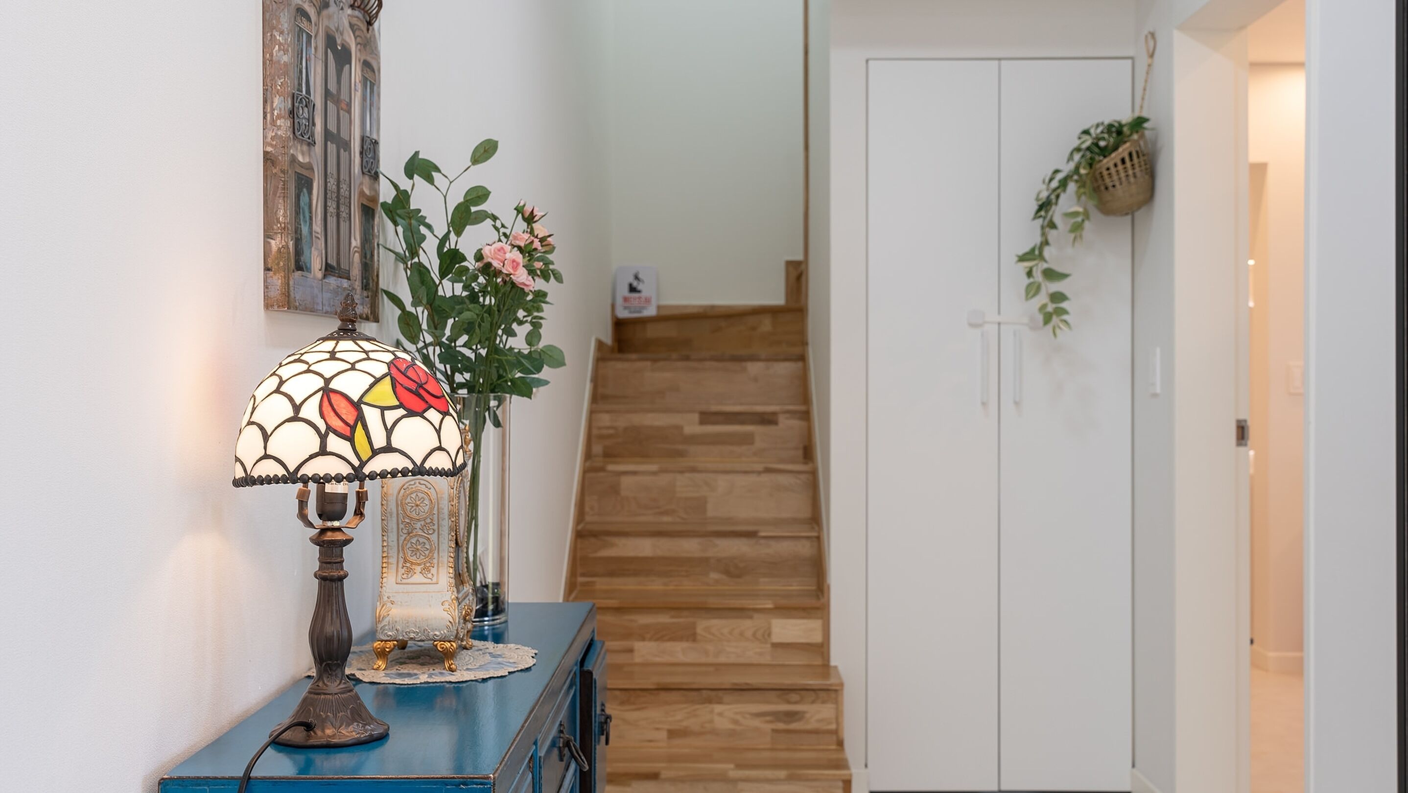A staircase with wooden steps leading to a white door, a blue cabinet with a decorative lamp, and a wall-mounted basket with greenery.