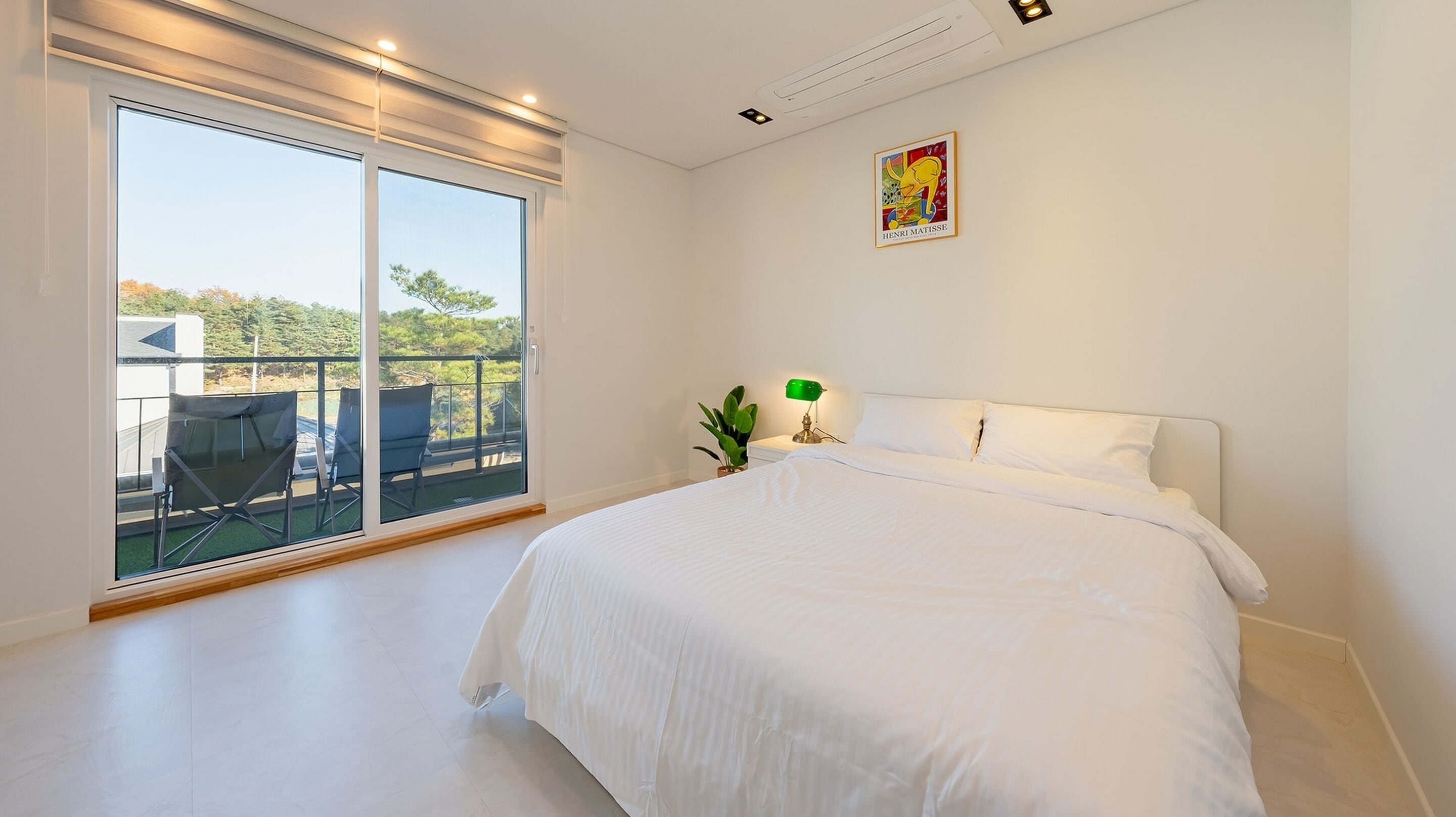 A bedroom with a large bed, a balcony with chairs, a green plant, and a painting on the wall.