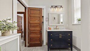 A bathroom with a wooden door, a dark vanity with gold handles, a white sink, and a mirror above it.