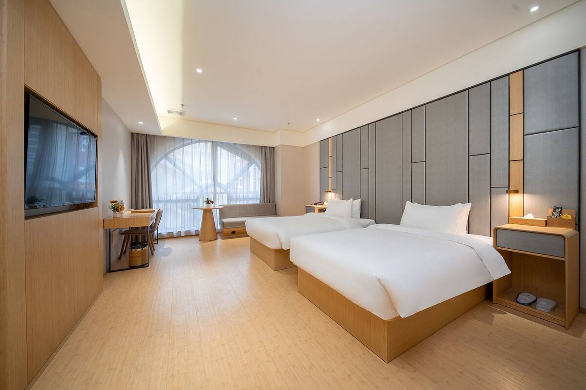 A modern hotel room with two beds, a desk, and a large window with sheer curtains.