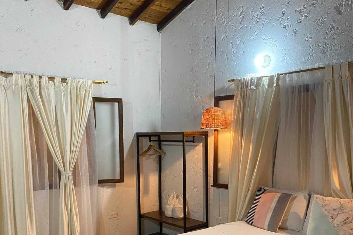 A bedroom with a bed, curtains, a hanging lamp, a shelf with a hanger, and a wall-mounted light.