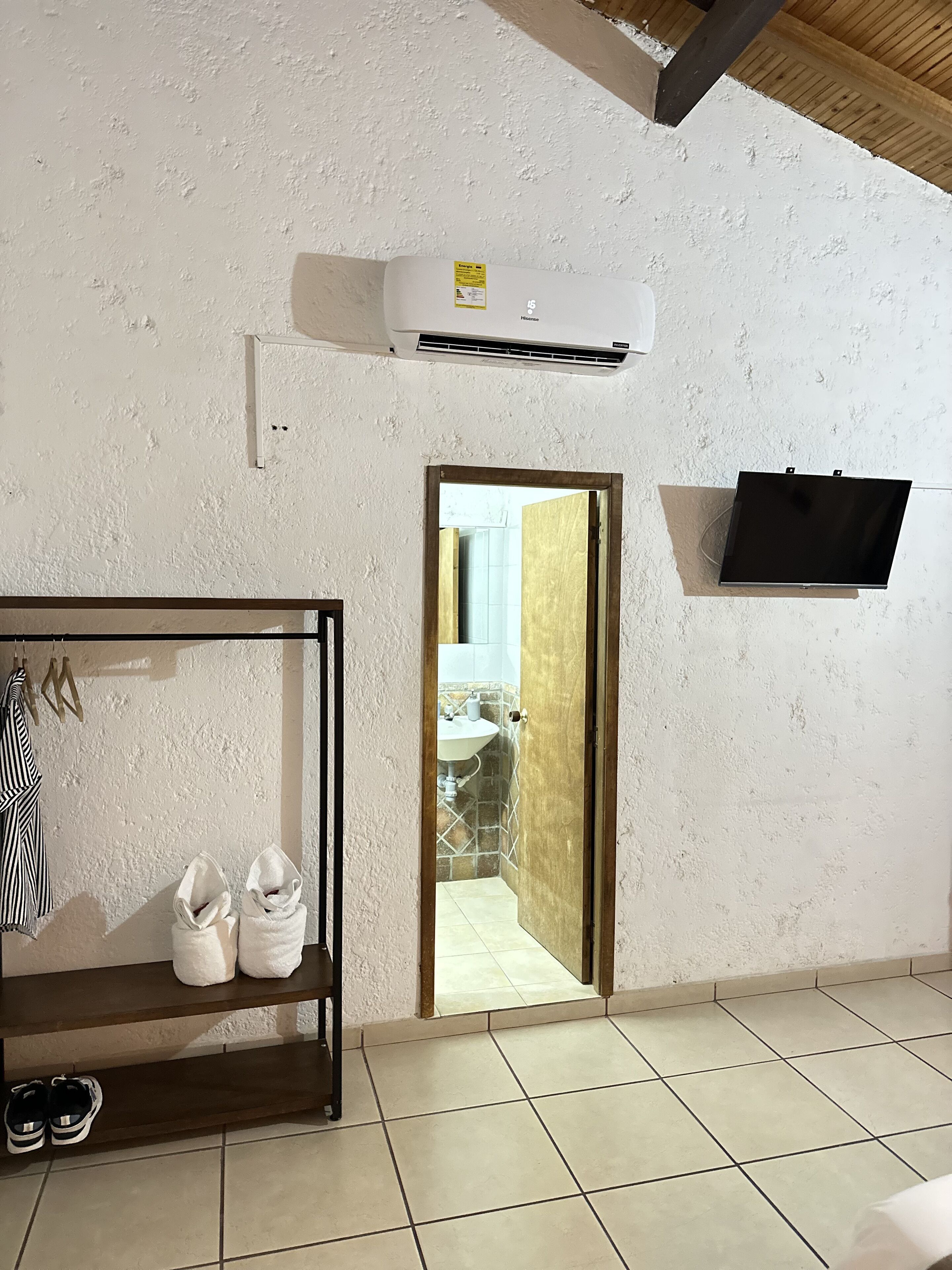 A room with a white textured wall, a mounted TV, a wooden shelf with towels and shoes, and a door leading to a bathroom.