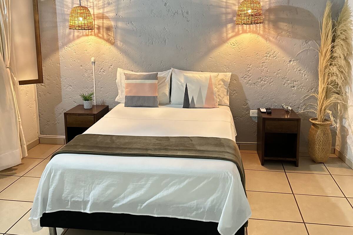 A bedroom with a bed, two bedside tables, a nightstand, a potted plant, and two hanging lamps.