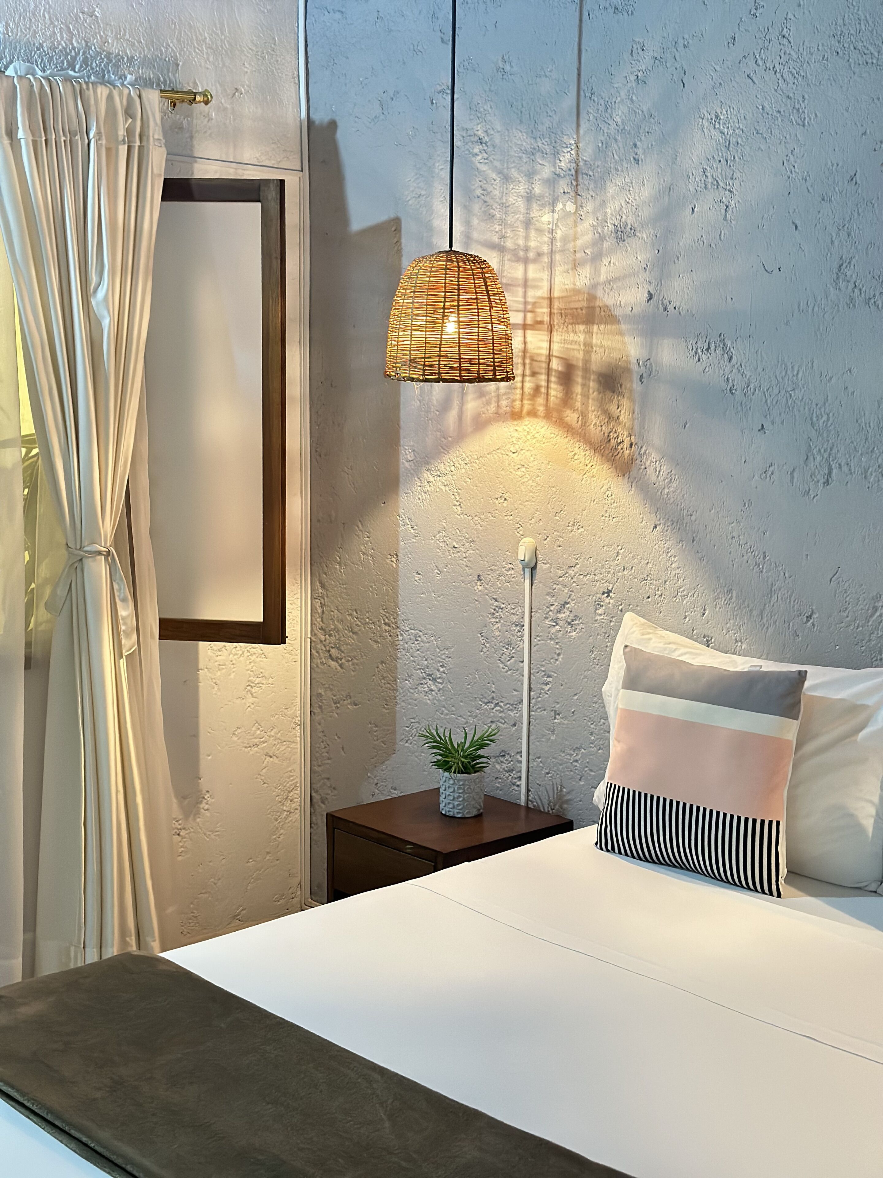 A bedroom with a bed, a bedside table with a lamp, a potted plant, and a curtain.