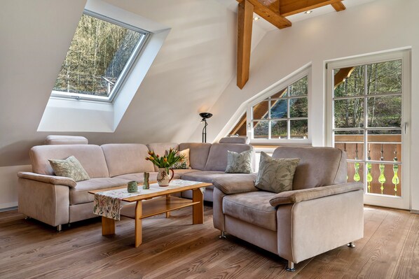 A loft-style room with a sloped ceiling, a sectional sofa, a wooden coffee table, and large windows offering a view of trees.