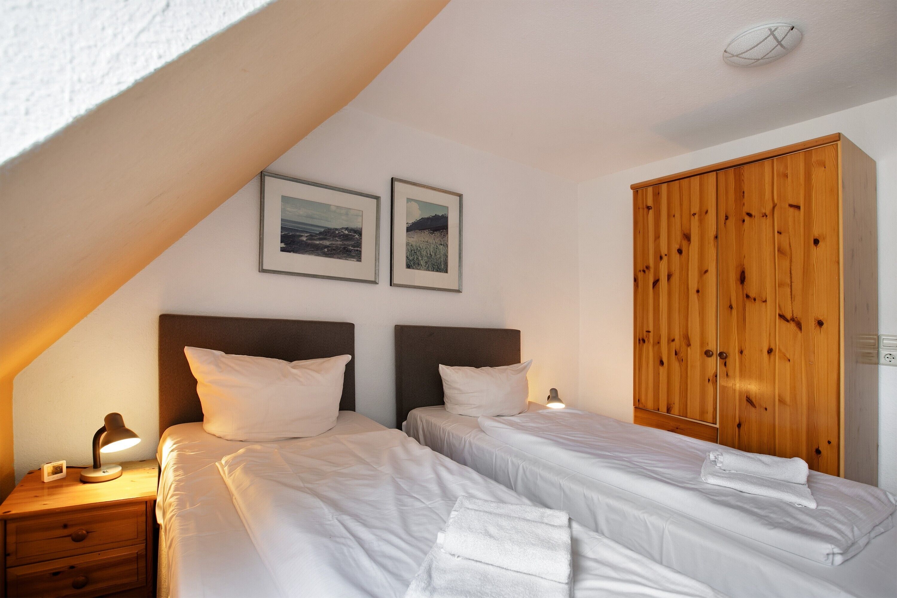A bedroom with two beds, a wooden wardrobe, and two framed pictures on the wall.
