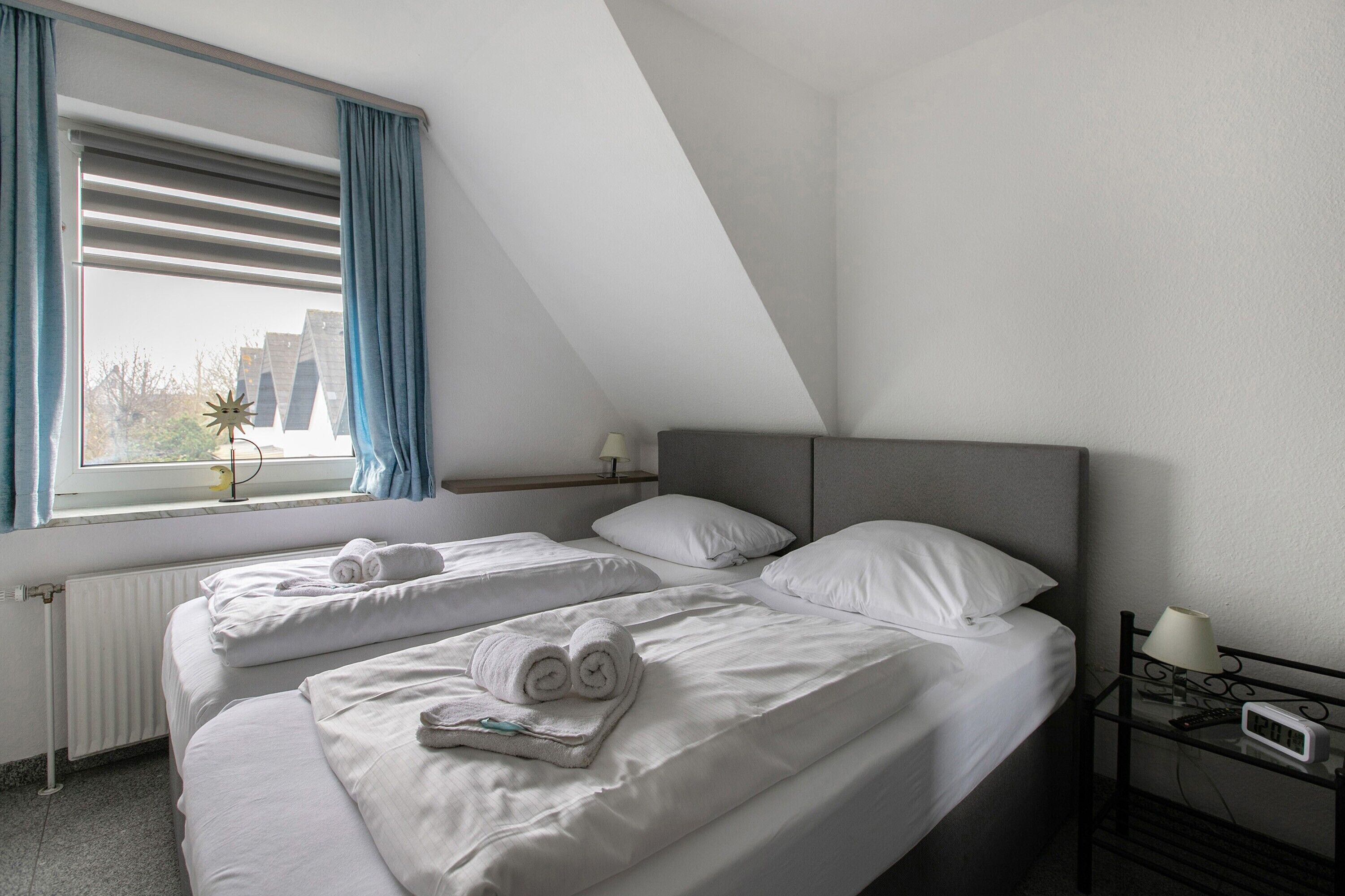 A hotel room with two beds, a window with curtains, a bedside table with a lamp, and a thermostat.