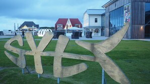 A wooden sign with the letters 'S' and 'X' on a grassy area, with buildings and a restaurant in the background.