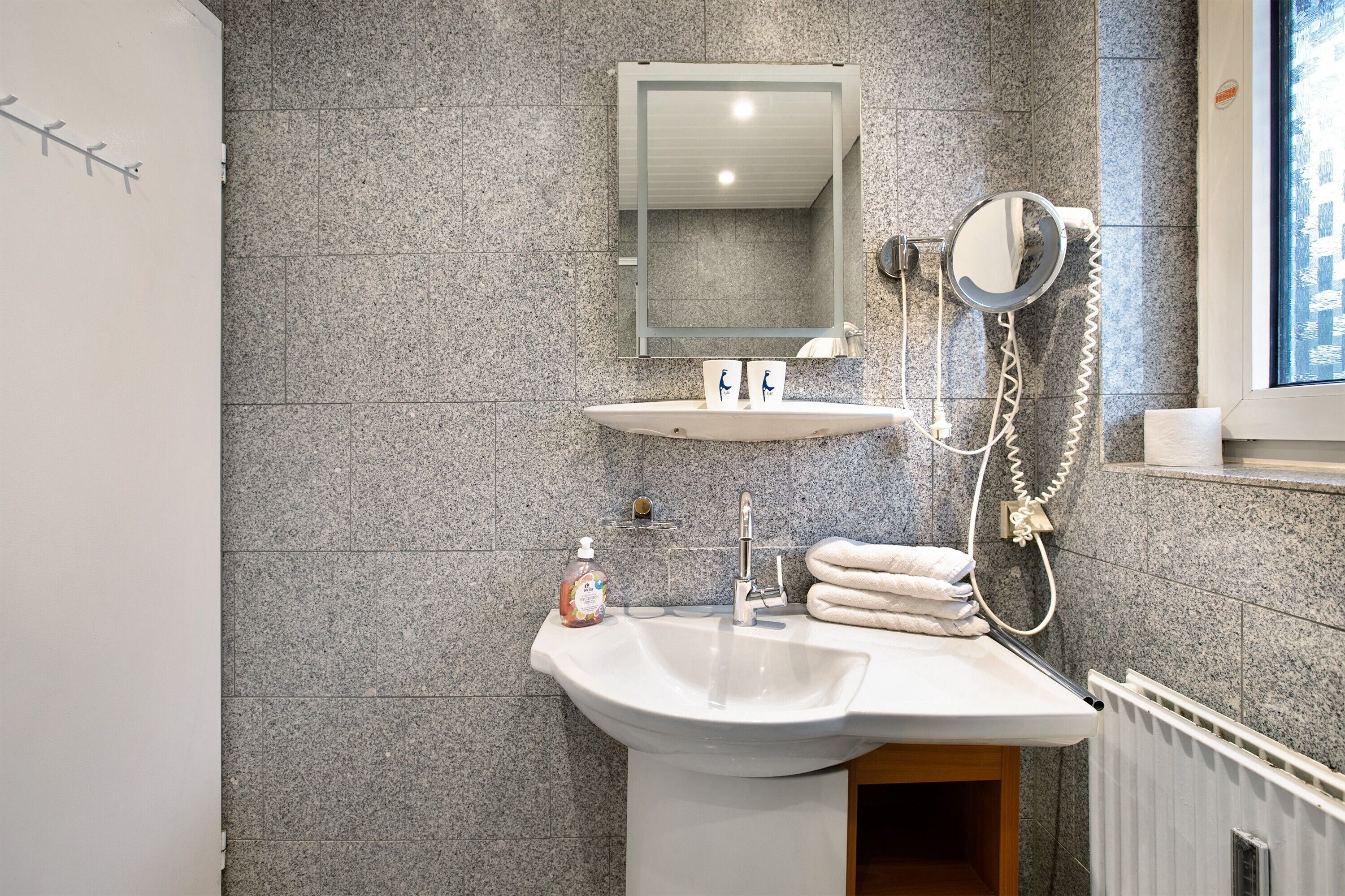 Une salle de bain avec un mur recouvert de carreaux de granit, un lavabo sur pied, un miroir, un porte-serviettes et un sèche-mains.