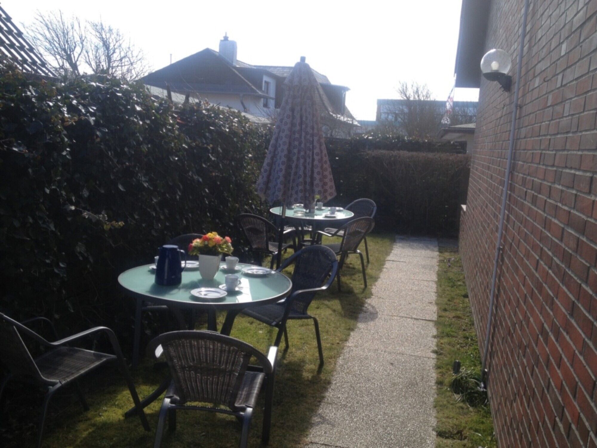 A patio with two round tables, chairs, a small table with a kettle and flowers, and a brick wall.