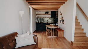 Ein gemütliches Wohnzimmer mit einem braunen Ledersofa, einem Holztisch und einem weißen Teppich. Eine Treppe führt zu einer Küche mit Holzböden und einer Rückwand im Fischgrätmuster.