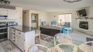 A modern kitchen with built-in appliances, a dining area with wicker chairs, a sofa, and a TV mounted on the wall. - Pacific View V484 (Gearhart)