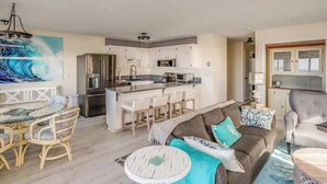 A modern kitchen with a central island, dining area with a glass table and chairs, and a living room with a sofa and armchairs. - Pacific View V484 (Gearhart)