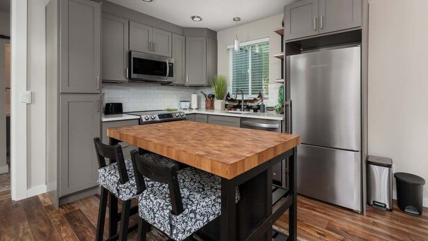 A modern kitchen with grey cabinets, a wooden island, stainless steel appliances, and black chairs with patterned cushions.