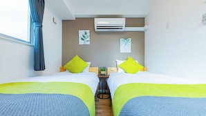 A hotel room with two beds, each with a green pillow, a small table with a plant, and an air conditioning unit on the wall. - JA Hotel Namba (Osaka)