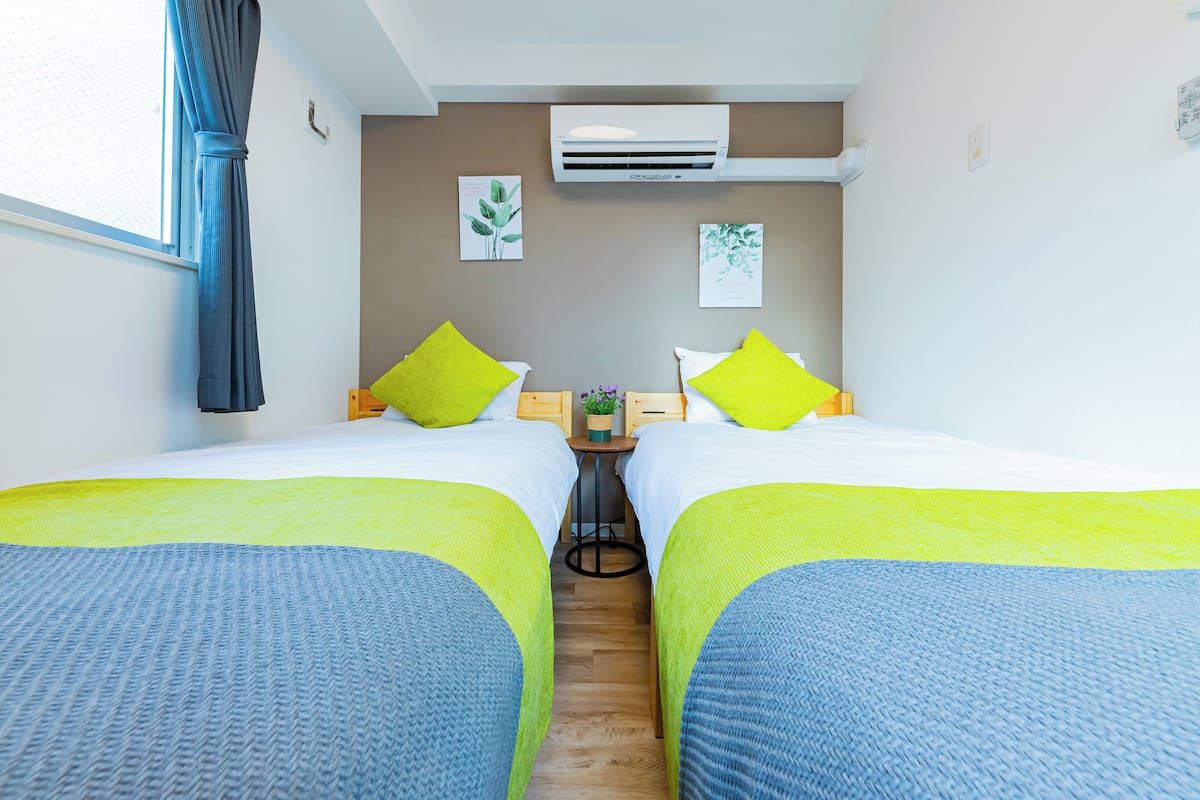 A hotel room with two beds, each with a green pillow, a small table with a plant, and an air conditioning unit on the wall.
