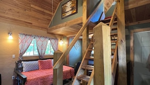 A rustic cabin interior with a wooden staircase, a bed with a red quilt, and a painting on the wall. - Clean cozy cabin within 1/2 mile of 2 lakes and 100’s of sq miles of wilderness. (West)