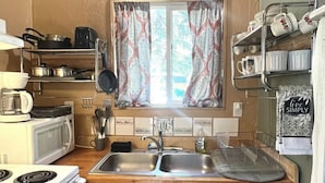 A compact kitchen with a stove, microwave, sink, and a window with patterned curtains. - Clean cozy cabin within 1/2 mile of 2 lakes and 100’s of sq miles of wilderness. (West)