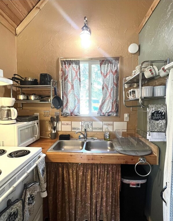 A compact kitchen with a stove, microwave, sink, and a window with patterned curtains.
